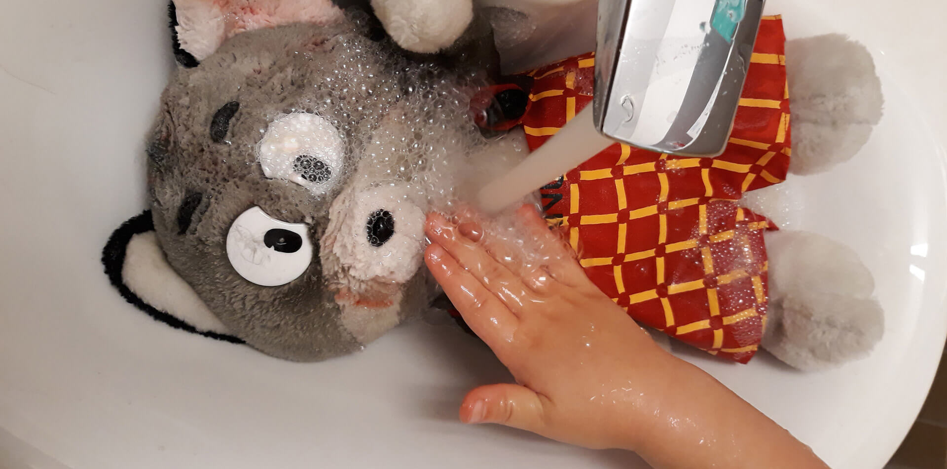 Kuvassa on käsienpesuallas, jossa makaa Manu-pehmolelu veden valuessa hanasta lelun päälle. Lapsen käsi pesee Manua, joka on selkeästi saanut punaista maalia kuonoon ja korvaan. Kuva: päiväkoti Ebeneser
