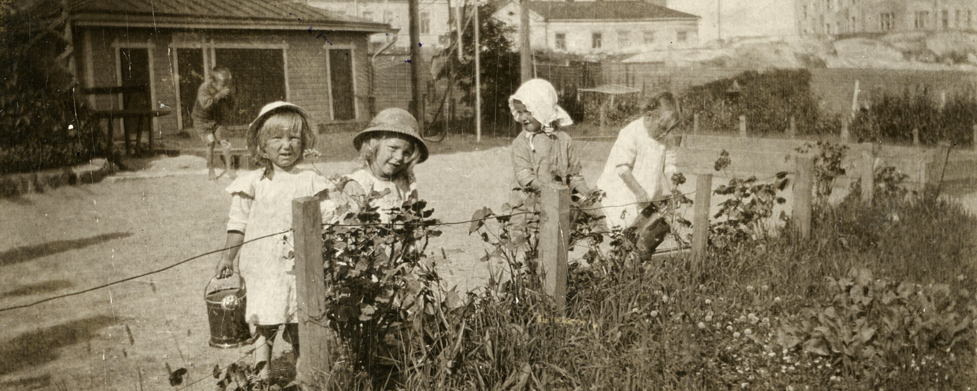 Kuvassa neljä lasta kastelemassa kukkia Ebeneserin pihalla. Taustalla on yksi lapsi. Taustalla näkyy Ebeneserin pihalla oleva puinen vaja sekä Helsinginkatu 1 tontin kalliot. Lastentarhamuseon valokuvakokoelma.
