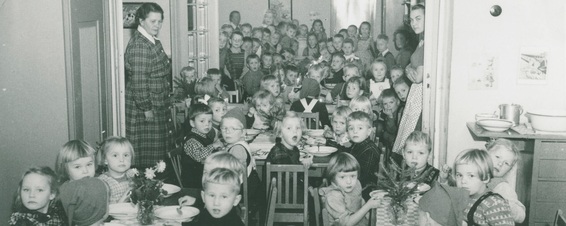 Rauhalan lastentarhan lapset yhteisellä joulupuurolla 1950-luvulla. Lastentarhassa oli lähes sen koko historian ajan tapana asetella jouluisin kaikki pöydät pitkän käytävän varrelle ikään kuin pitkäksi yhteiseksi pöydäksi. Pöydillä on kukkia, kuusen havuja ja kynttilöitä. Osalla lapsista on tonttulakki päässä.