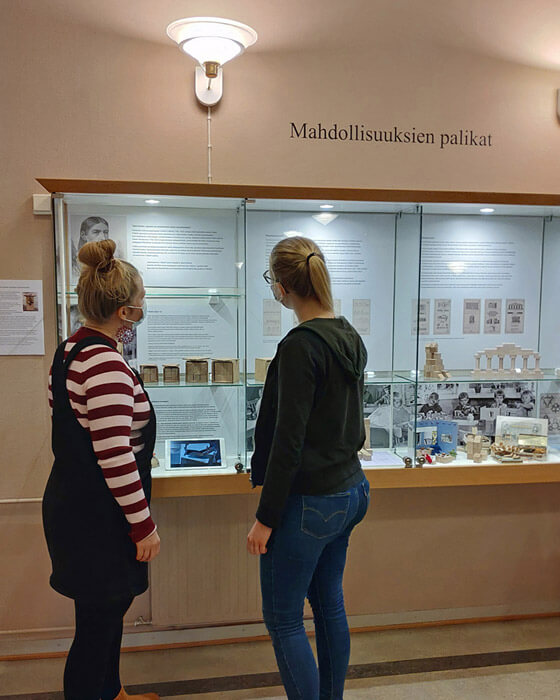 Kuvassa kaksi opiskelijaa seisovat seinälle kiinnitetyn vitriinin ääressä ja katselevat sen sisältöä. Vitriinissä on esillä Mahdollisuuksien palikat näyttely, joka esittelee Friedrich Fröbelin leikkilahjat nro 1-6 sekä niiden eri käyttötarkoitukset.