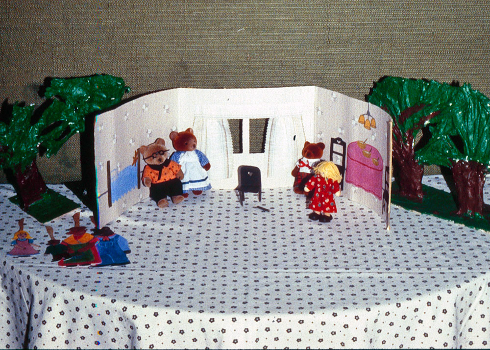 Kuvassa 1970-luvun päiväkodin pöyäteatterihetki. Pieni sermi kuvastaa taloa, jonka ympärille on aseteltu askarreltuja puita. Sermin sisäpuolella, talossa, on kolme karhuhahmoa ja Kultakutri. Yksityiskokoelma.