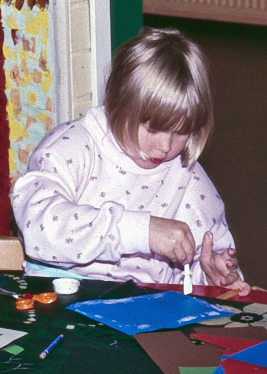 Lapsi levittää liimaa keskittyneesti liimatikkua apunaan käyttäen siniselle kartongille. Yksityiskokoelma.