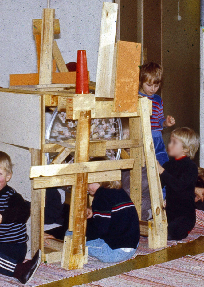 Kuvassa raakalaudoista päiväkodin sisätiloissa rakennettu rakennelma. Lapsia on touhuamassa rakennelman jokaisella puolella ja osa sen sisällä. Yksityiskokoelma