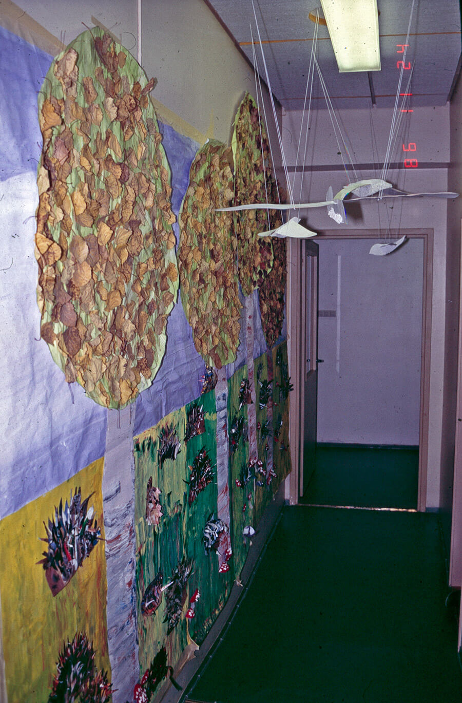 Kuvassa lapsiryhmän tekemä, seinälle kiinnitetty virikealusta, jonka edessä lentää paperista tehty, naruilla kattoon kiinnitetty lintu. Yksityiskokoelma.