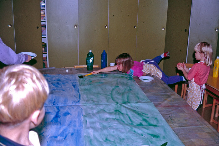 Kuvassa lapset maalaavat suurta, koko pöytäpinnan kokoista paperia sinertävällä ja vihertävällä värillä. Yksityiskokoelma.