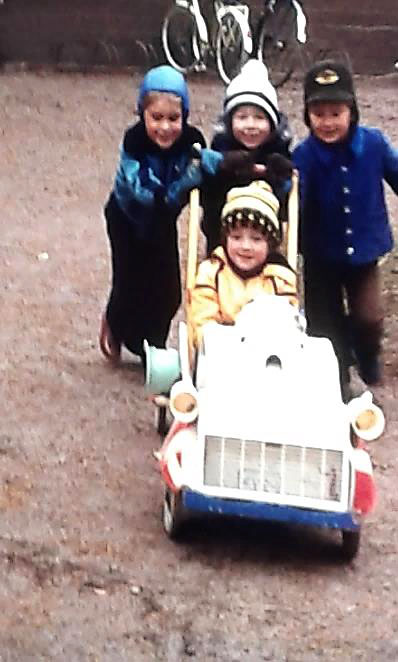Kuvassa on neljä poikaa leikkimässä pihalla työnnettävällä autolla, ohjaajan kädet ovat ratissa nopeusmittarin takana ja kolme kaveria työntää takaa turvakaaren tapaisesta rakenteesta. Yksityiskokoelma.
