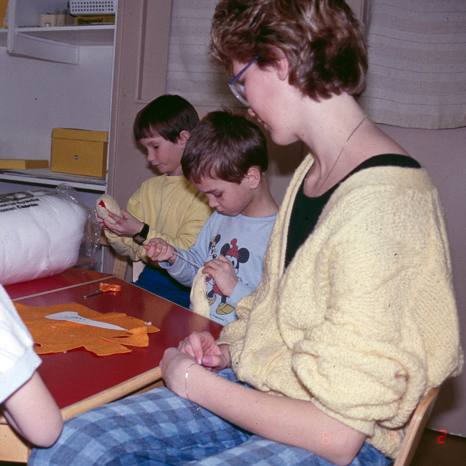 Kuvassa punapintainen tyypillinen 1970-80-luvun päiväkodin pöytä, jonka ääressä lapset askartelevat ilmeisesti kanoja tai tipuja. Yksi lapsi ompelee huopaista keltaista hahmoa ja toinen lapsi täyttää omaa hahmoaan vanulla. Aikuinen katsoo lasten toimintaa vieressä. Pöydällä on oranssia huopaa sekä sakset. Yksityiskokoelma.