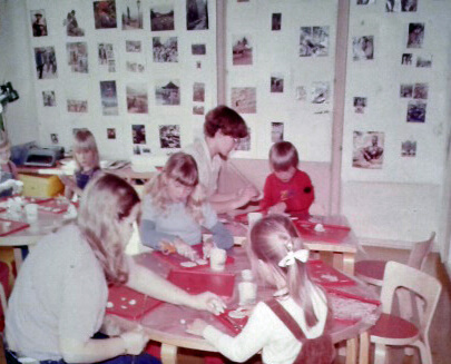 Kuvassa lapsia ja aikuisia ilmeisesti muovailemassa massasta pieniä figuureja tai muotoja. Pöydät ovat Alvar Aallon tuotantoa - puiset ja tasoiltaan punapintaiset. Taustalla seinällä ilmeisesti lehdistä leikattuja kuvia. Yksityiskokoelma.