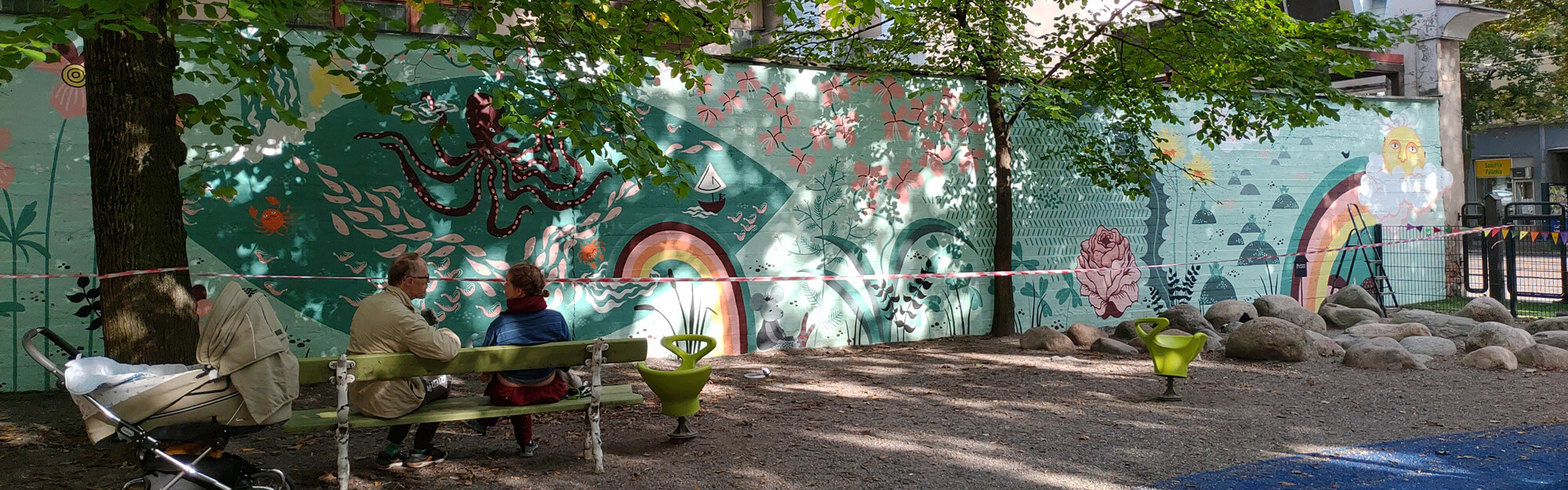 Taiteilija Essi Ruuskanen istumassa maalaamansa muraalin edessä Lastentarhamuseon leikkipihalla. Lastentarhamuseo.
