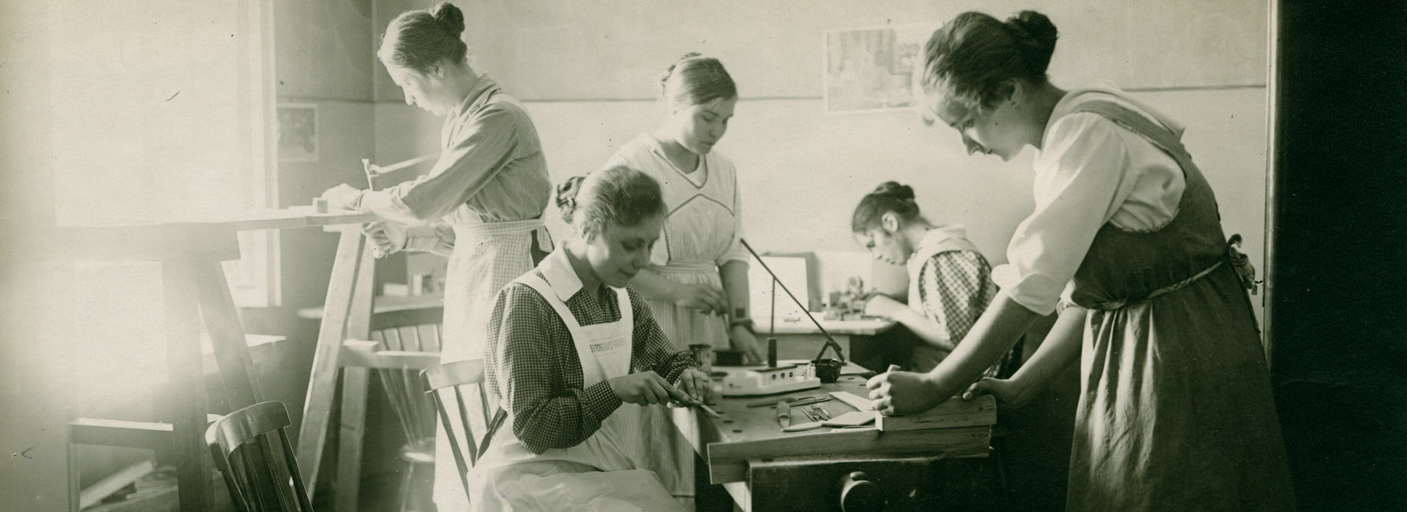 Kuvassa viisi lastentarhanopettajaopiskelijaa 1910-luvulla veistotunnilla Ebeneserin seminaarissa. Osa opiskelijoista seisoo ja osa istuu höyläpenkin äärellä työstäen tarkasti erilaisia puutöitä. Lastentarhamuseon valokuvakokoelma.
