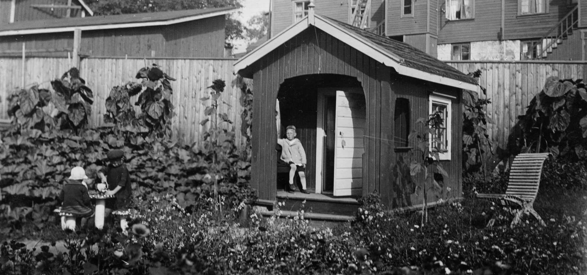 Kuvassa Ebeneser-talon pihamaata 1920-luvulla. Vehreiden kukkaistutusten taustalla näkyy leikkimökki, jonka kuistilla istuu lapsi tuolilla. Vieressä kaksi lasta leikkii kärpässienituolin ja -pöydän luona. Pitkiksi kasvaneet auringonkukat nojaavat leikkimökin takana olevaa Ebeneserin pihaa rajaavaa puuaitaa vasten. Aidan takana on kaksikerroksisia puisia asuintaloja. Lastentarhamuseon valokuvakokoelma.