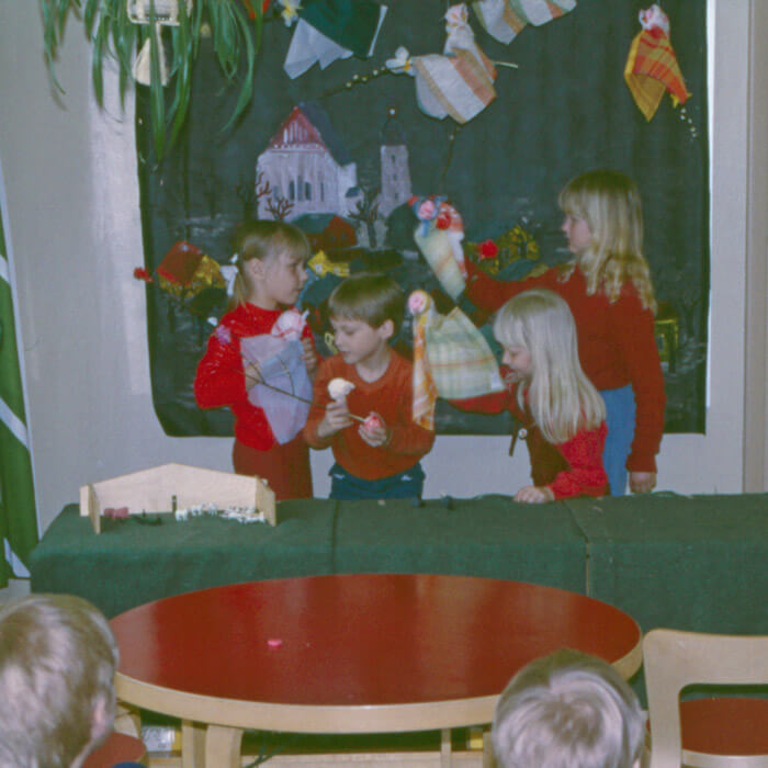 Neljä lasta ovat asettuneet pöydän taakse esittämään pöytäteatteria. Lapsilla on kädessään hahmoja ja pöydälle heidän eteensä on levitetty vihreä liina, ikään kuin ruohikoksi. Pöydällä on jonkinlainen aitaus, jonka sisällä on eläimiä. Lasten takana seinällä on maisemamaalaus ja katosta lasten yllä roikkuu pääsiäisnoitia, jotka lentävät pajunkissoja kasvavilla pajun oksilla. Lastentarhamuseon valokuvakokoelma.