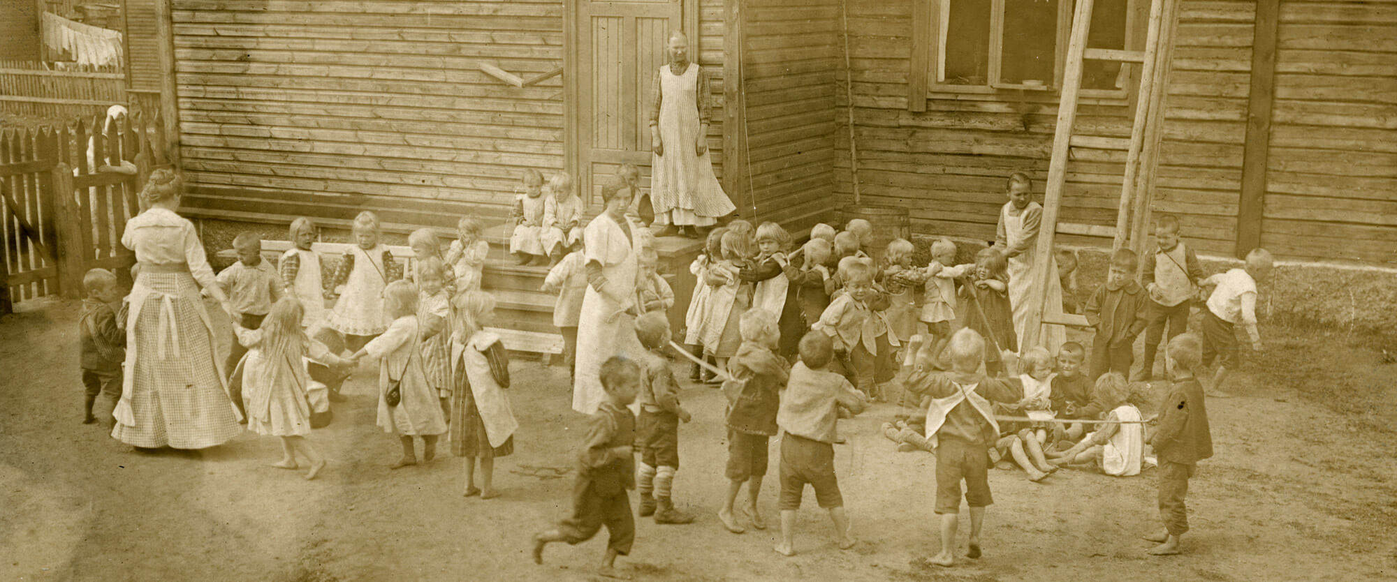 Sörnäisten kansanlastentarhan lapsia ja lastentarhanopettajia leikkimässä Suruttomain huvilan edessä noin vuonna 1890. Osa lapsista leikkii piirileikkiä, osa kulkee jonossa kädet toistensa harteilla, osa pitää kiinni pitkästä köydestä. Suurimmalla osalla lapsista on esiliinat yllään ja osalla heistä on olan yli menevä pieni laukku. Suurin osa lapsista on paljain jaloin. Vasemmalla olevan puisen aidan takaa katsoo kaksi ihmistä, toinen luultavasti lapsi ja toinen aikuinen. Vasemmalla talojen välissä on pyykit kuivumassa pyykkinarulla.