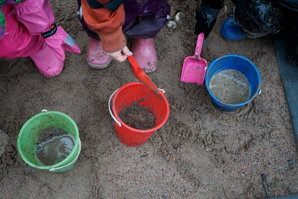 Kuvassa on kolme eriväristä pientä muovisankoa, joiden jokaisen ääreelle on kumartuneena lapsi rajautuen kuitenkin kuvasta pois. Lasten kädessä on pienet värikkäät lapiot, joilla he oletettavasti lastaavat ämpäreihin kuraista maata.