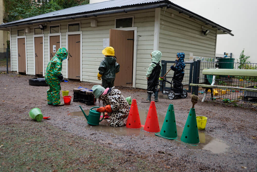 Kuvassa on viisi lasta kuravaatteissaan pihaleikeissä. Kuvan taustalla on puinen vihreä rakennus.