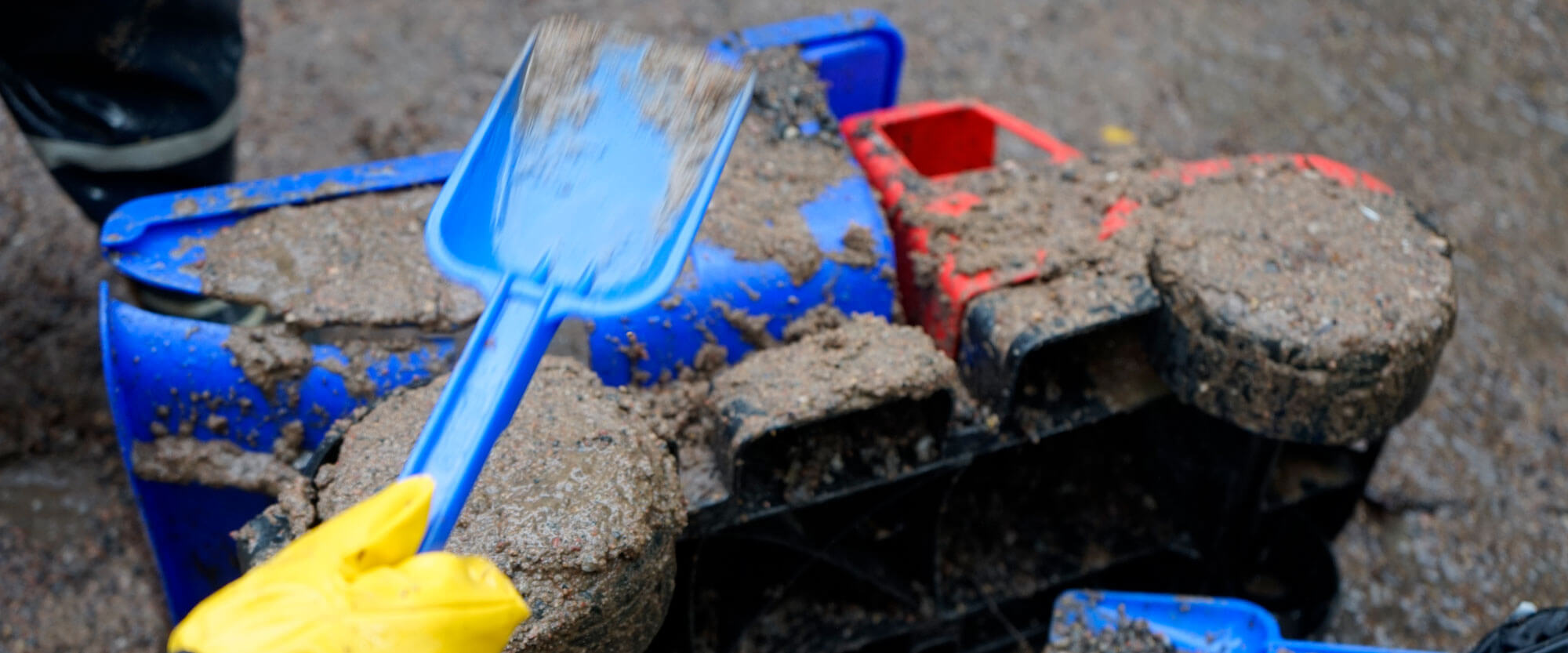 Kuvassa on muovinen lelukuorma-auto kyljellään makaamassa kuraisessa maassa. Auton päälle on levitetty kuraa ja kuvaan on rajautunut kaksi sinistä lasten muovilapiota, jotka ovat ilmassa auton päällä.