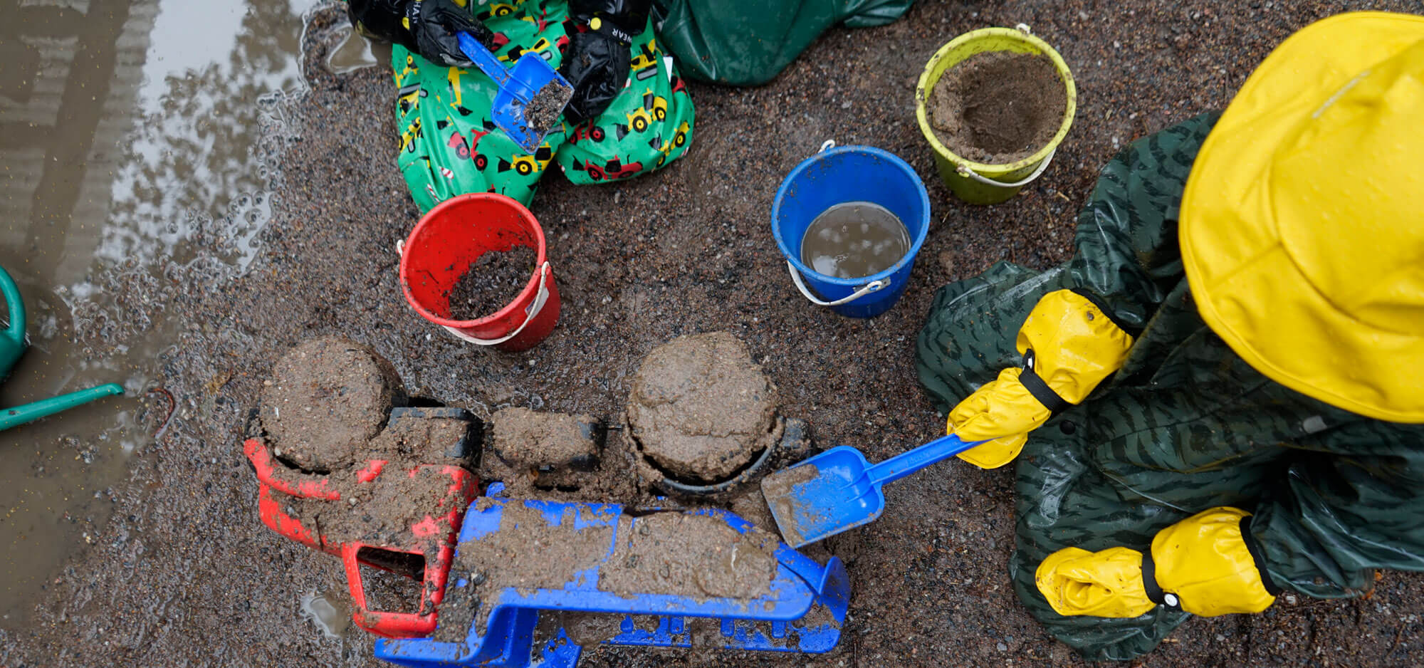Kuvassa on kaksi lasta sadevaatteet päällä istumassa kuraisessa maassa. Lasten vieressä on muovinen kuorma-auto, jonka päälle toinen lapsi levittää kuraa. Lasten vieressä on kolme värikästä pientä muoviämpäriä, joissa on kuraa.