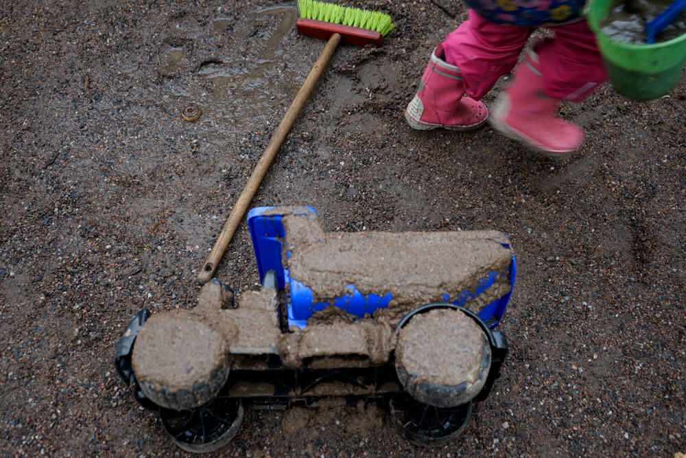 Kuvassa on muovinen lelukuorma-auto kyljellään makaamassa kuraisessa maassa. Auton päälle on levitetty kuraa ja sen vieressä on lasten katuharja sekä kumppareihin pukeutuneen lapsen jalat. Lapsi on ilmeisen kiireisenä poistumassa auton luota kantaen vihreää ämpäriä täynnä kuraa.