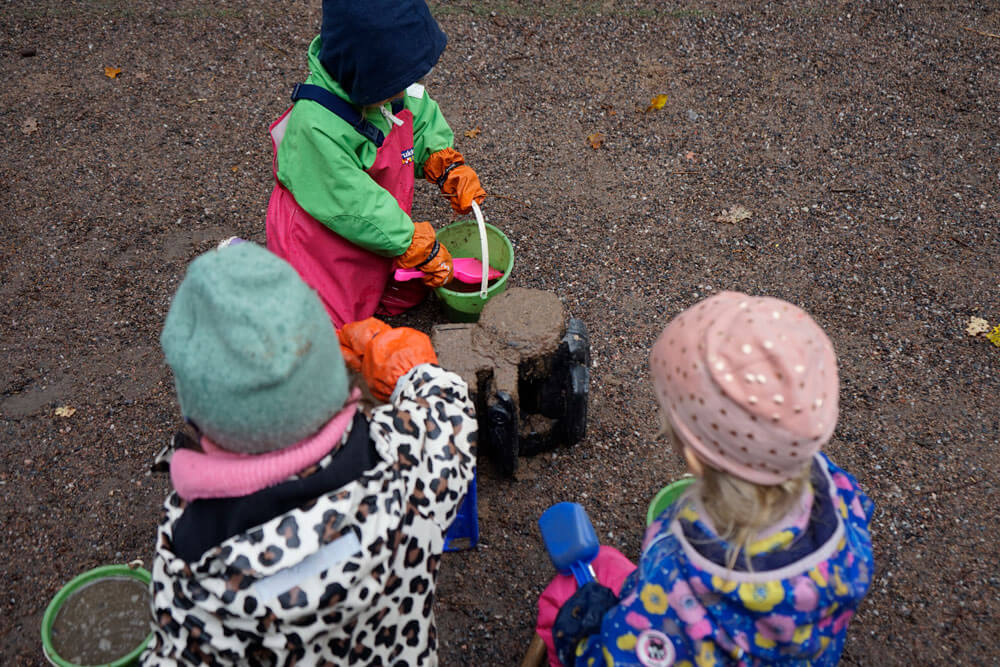 Kuvassa on kolme lasta sadevaatteisiin pukeutuneina muovisen kuorma-auton äärellä istuvina. Lapsilla on kädessään muoviset lapiot, joilla he laittavat kuraa kuorma-auton päälle.
