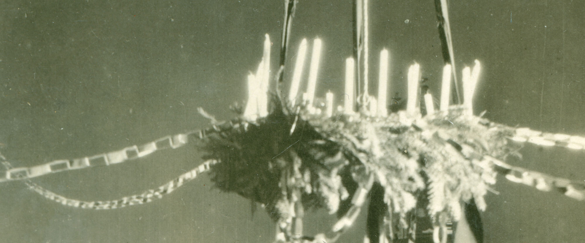 Lastentarha Barnabon (perustettu 1913) lapsia istuu kahdessa rivissä penkillä kuvan alareunan myötäisesti. Lasten takana on seinä, jonne on aseteltu kuvia tai piirustuksia tasaisin välimatkoin. Suuressa osaa kuvassa on adventtiseppele, joka roikkuu katosta nauhoin. Seppele on tehty aidoista havuista, koristeltu paperiköynnöksin ja aidoin kynttilöin, Kuva otettu arviolta vuosien 1913-1919 välillä. Lastentarhamuseon valokuvakokoelma.