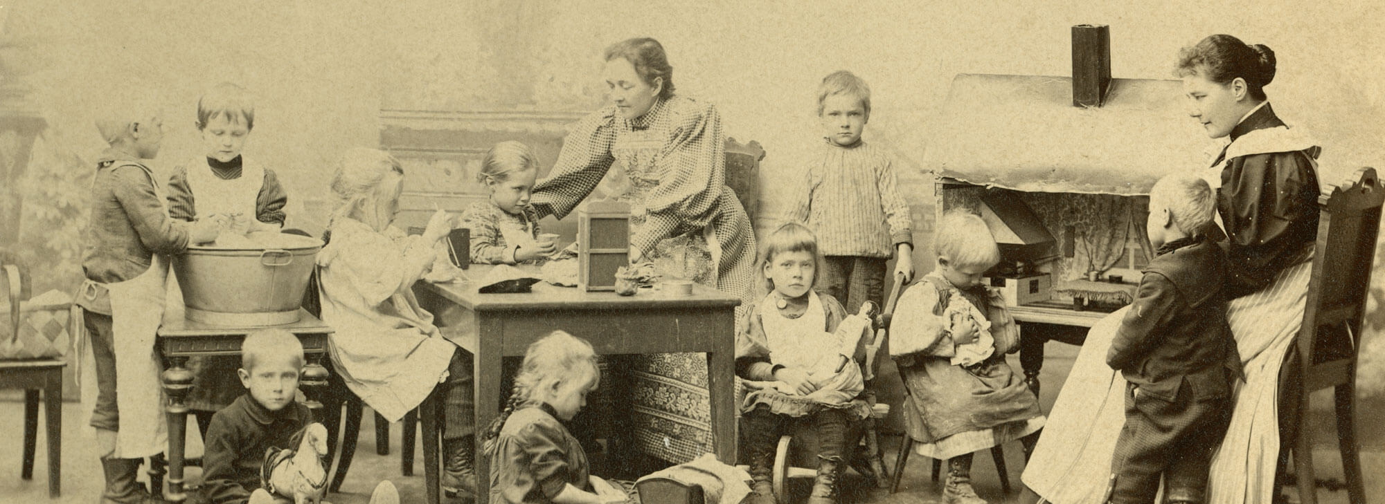 Kuvassa Sörnäisten lastentarhan lapsia touhuissaan Suruttomien huvilassa, Alli Tryggin rakennuttamssa Kansankodissa. Kuvassa on 10 lasta ja kaksi aikuista. Aikuiset ovat suomalaisen varhaiskasvatuksen uranuurtajat ja "äidit" Hanna Rothman sekä Elisabeth Alander. Kuvassa osa lapsista leikkii ja osa tekee nk. työtehtäviä, kuten neuvolavat ja pesevät nukenpyykkiä. Rothman ja Alander ovat yhdessä lasten kanssa touhuilemassa. Lastentarhamuseon valokuvakokoelma