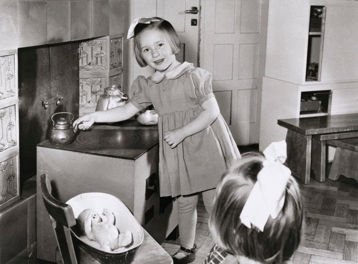 Päiväkoti Kyläneva, rusettipäinen lapsi leikkikeittiössä keittämässä kahvia. Etualalla vauvanukke vaunuissaan. Helsingin kaupunginmuseo