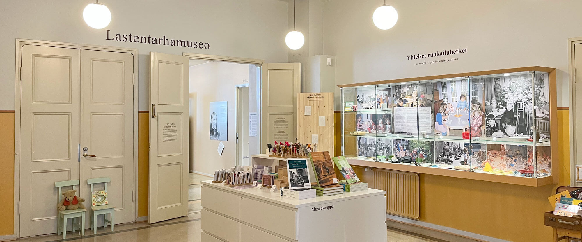 Näkymä kolmannen kerroksen saavuttaessa kolmannen kerroksen aulatilaan. Aula avautuu valoisana. Seinien alaosat on maalattu lämpimän keltaisella ja yläosat vaaleanharmaalla. Saliin johtavat pariovet ovat avoinna kuvan taustalla ja niiden vieressä sijaitsee seinälle kiinnitetty lasivitriini. Keskellä huonetta on valkoisille senkeille rakennettu esittelypöytä museokauppatuotteille. Katosta roikkuvissa pallovalaisimissa on lämmin hehku.