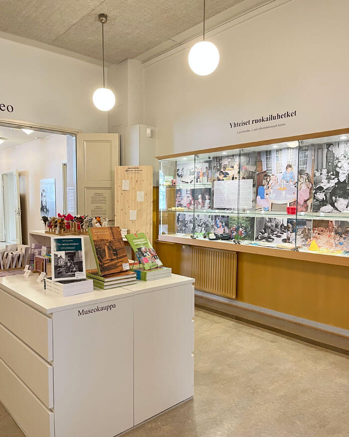 Näkymä porrastasanteen ovelta kohti seinävitriiniä. Etualalla kahdesta valkoisesta senkistä koostuva esittelytaso museokauppatuotteille. Seinälle jäänyt vanha vitriini sopii huoneen remontoituun ilmeeseen. Taustalla pariovet avautuvat kohti valoisaa Lastentarhamuseon salia.