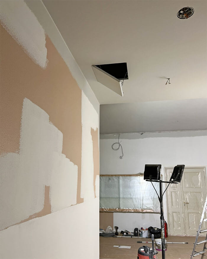 Aulan ja siitä johtavan käytävän seinät on tasoitettu ja paikkomaalattu. Katon huoltoluukku on avoinna. Työmaavalo valaisee remontoitavaa tilaa.