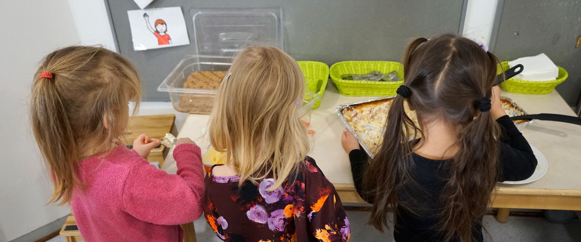 Yhteiset ruokailuhetket - päiväkoti- ja lastentarhamuistojen keruu -näyttelyjulisteessa kolme lasta seisoo selin kameraan ja ottavat ruokaa tarjoilupöydältä.