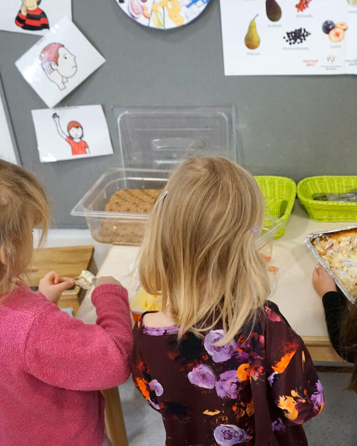 Yhteiset ruokailuhetket - päiväkoti- ja lastentarhamuistojen keruu -näyttelyjulisteessa kolme lasta seisoo selin kameraan ja ottavat ruokaa tarjoilupöydältä.