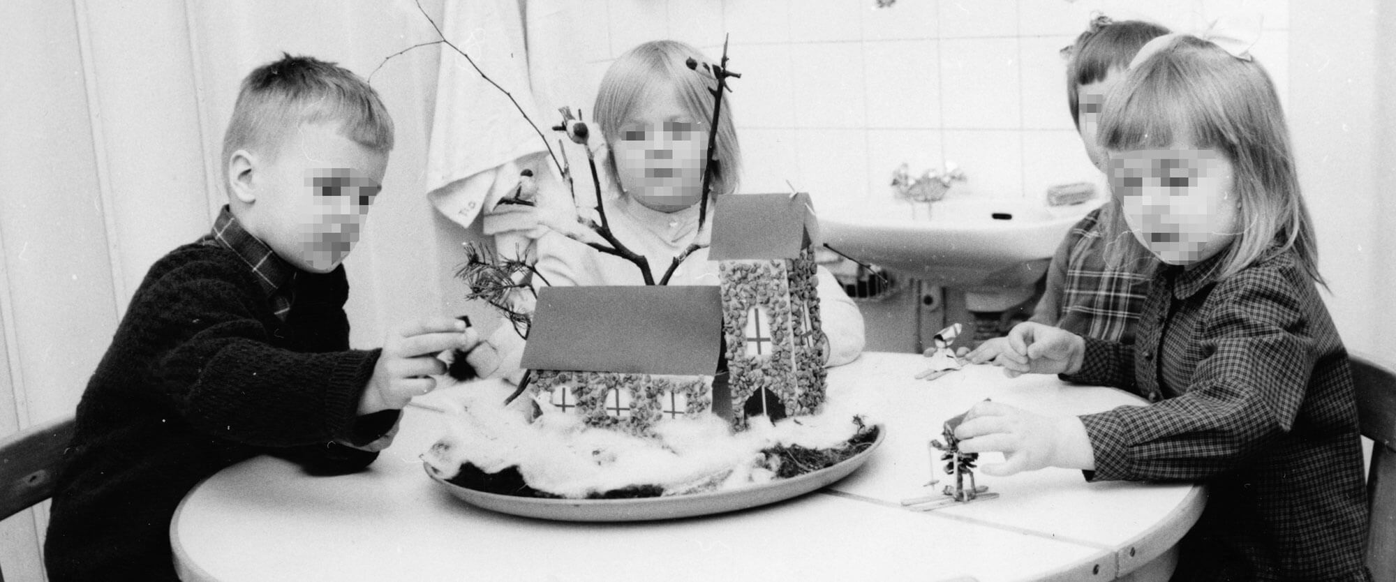 Neljä lasta istuu mustavalkoisessa kuvassa pöydän äärellä ja tutkivat pöydälle asetettua kirkon pienoismallia. Kirkko on asetettu tarjottimelle, jolla on oletettavasti pumpulia, joka demonstroi lunta. Yksi lapsista liuvuttaa pöydän pintaa pitkin käpytonttua, jolla on pienet leikkisukset jalassa.